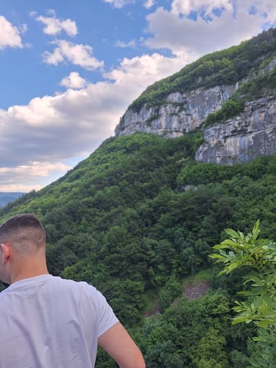 The image captures a scene in Zasele, Bulgaria, featuring a man standing on an elevated area with a backdrop of a tall, rocky cliff face covered in lush green vegetation. The cliff also displays geological features like sedimentary layers. The sky above is partly cloudy with blue patches. The man is wearing a white t-shirt, and only a portion of his head and shoulder is visible. A few green branches with leaves extend from the vegetation towards the upper right corner of the frame.