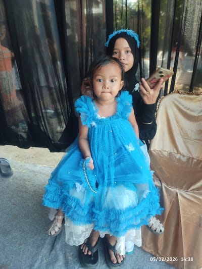 A young child in a frilly blue dress sits on someone's lap, holding a pearl necklace. An older girl in a black hijab and matching blue headband is behind, looking at a cell phone with a floral case. The background includes a dark curtain, a window, and a gold-covered chair. Sandals and flip-flops are visible on the floor. The date "05/02/2026" is stamped at the bottom right of the image.