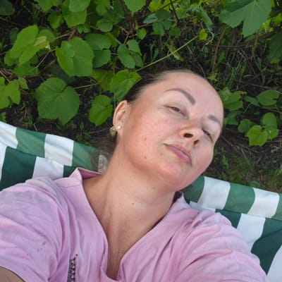 A woman with fair skin and brown hair is lying down on a striped green and white surface, outdoors. She is wearing a pink shirt. Her head is tilted to the side, and she is looking upwards. Green foliage is visible in the background, suggesting a natural, possibly wooded environment. The scene is likely captured in Novoostrovka, Russia, given the context provided.
