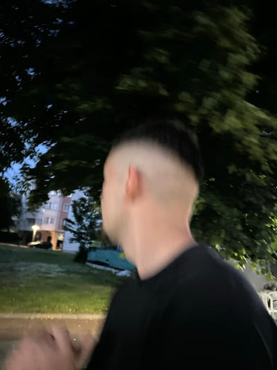 The image depicts a twilight or nocturnal outdoor scene in Dzhebel, Bulgaria. A single individual, observed from the left posterior aspect, occupies the foreground. This person exhibits a high skin fade haircut on the left side, transitioning from a closely shaved area near the ear to darker, longer hair superiorly. They are wearing a plain, dark-colored, short-sleeved garment. The left arm and hand are visible in the lower-left quadrant, exhibiting significant motion blur, suggesting movement or camera shake.
In the mid-ground and background, dense foliage from large trees with dark green leaves dominates the upper and right portions of the frame. Patches of a dim, blue-grey sky are discernible through the tree canopy. A residential building, multiple stories high with illuminated windows, is partially visible in the left background. A prominent streetlight with a warm yellow-orange glow illuminates a portion of the grassy ground and adjacent structures in the mid-left. The overall scene is dimly lit, with artificial light sources providing illumination. The image quality is affected by motion blur and low light conditions.