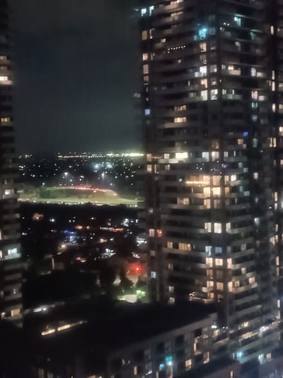 The image depicts a nighttime cityscape, likely from a high vantage point, showcasing illuminated high-rise buildings and a distant urban landscape. The perspective suggests the viewer is situated within a building, looking out over the city.
In the foreground, to the left and right, are portions of two large, modern apartment or condominium buildings. These structures are characterized by numerous windows, many of which are brightly lit, suggesting occupancy within the units. The architectural style appears to be contemporary, with clean lines and repetitive window patterns.
The middle ground reveals a vast expanse of the city at night. Numerous smaller buildings and houses are visible, each contributing to the overall illumination with their own lights. A prominent feature in this area is a wide, multi-lane road, likely a highway or major arterial street, with visible streaks of light indicating moving vehicles. Streetlights are also scattered throughout the landscape, casting pools of light.
In the distance, the urban sprawl extends towards the horizon, marked by a faint glow of lights against the dark night sky. The overall impression is one of a densely populated and active urban center.
The location of Mississauga, Canada, is identifiable through the specific architectural style of the buildings, the layout of the roads, and the general character of the urban development, which is consistent with the suburban and commercial areas of Mississauga. The presence of distinct high-rise residential towers and the surrounding infrastructure aligns with known developments in the city, particularly in areas like Square One or along the Hurontario corridor.