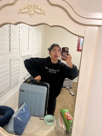 A young woman is taking a mirror selfie. She is wearing a black hoodie and glasses. Her tongue is sticking out. She is holding a phone in her right hand. Next to her is a light blue suitcase. Behind her is a white shuttered door. In the background is a bed, a gray chair, and a pair of shoes. There is also a bag of chips and a pillow on a shelf. The location cannot be determined.