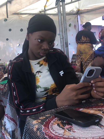 A young woman with dark hair covered by a black headscarf is holding two smartphones. She's wearing a black Fila jacket over a white sunflower-print shirt. The woman is sitting at a table covered with a patterned tablecloth. Another person, wearing a yellow and black dress, is sitting nearby. The scene appears to be outdoors, under a white tent-like structure. The location is Nteje, Nigeria.