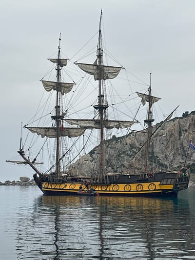 The image features a majestic multi-masted sailing ship, reminiscent of a historical tall ship or galleon, calmly floating on water against a backdrop of a rugged, light gray rocky cliff face. The ship's hull is predominantly dark, but it is accentuated by a striking yellow stripe that runs along its side. This yellow stripe is adorned with several dark, circular emblems, many of which clearly display the Roman numeral "II".
The vessel boasts at least three prominent masts, each fitted with numerous furled sails and an intricate network of ropes and rigging. Several small human figures are discernible on the ship's deck, particularly near the stern, and possibly among the rigging.
The calm water in the foreground creates distinct reflections of both the ship's hull and the textured cliff face. The sky above is uniformly overcast and light gray, casting a soft, diffused light over the entire scene. A small flag, possibly a Union Jack or similar, can be seen flying near the ship's stern, atop one of its masts. Faintly visible on the ship's stern, above a row of windows, is the text "MARTELLOO".
The scene suggests the ship is either anchored or docked close to the shore in a bay or harbor. The peaceful coastal setting, with its dramatic cliff, aligns with locations such as Datça, Turkey, as provided in the context. The ship appears to be either a well-maintained historical vessel or a meticulously crafted replica.