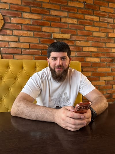 A man with a beard and dark hair is sitting at a dark wooden table. He is wearing a white t-shirt with a graphic design. He is holding a rose gold smartphone in his right hand and has a black smartwatch on his left wrist. The man is sitting on a yellow tufted couch. Behind him is a brick wall with orange and brown bricks. The lighting suggests it is daytime. The man's expression appears neutral.