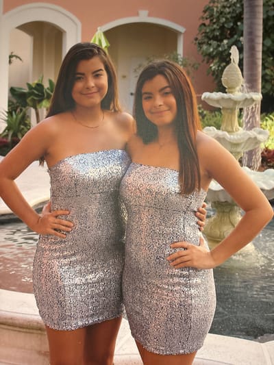 Two young women, likely twins, pose for a photograph outdoors in front of a water fountain and a stucco building with arched openings. Both are wearing strapless, silver sequined dresses that reach mid-thigh. The woman on the left has her left hand on her hip and her right hand slightly in front of her waist. The woman on the right has her left hand resting on the hip of the woman on the left, and her right hand is near her own hip. Both have long, dark brown hair and are smiling at the camera. The background suggests a resort or a manicured garden area, possibly near a body of water. The scene is well-lit, indicating daytime.