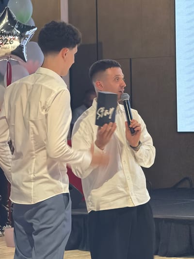 Two young men are on a stage in front of an audience, though the audience is not fully visible. The man in the foreground is wearing a white shirt and dark blue pants. He has dark curly hair and is looking up. He is surrounded by balloons, including a silver star-shaped balloon with text on it. The man in the background is also wearing a white shirt and dark pants. He is holding a microphone in his right hand and a black rectangular object in his left hand. He is speaking into the microphone and looking towards the audience. A large screen is visible above the stage.

The star-shaped balloon has the Cyrillic text "Випуск 2026" which translates to "Graduation 2026", and "ліцей №1", meaning "Lyceum No. 1". The event appears to be a graduation ceremony for Lyceum No. 1 in Khotianivka, Ukraine. The lighting suggests an indoor event, possibly in the evening. The atmosphere seems celebratory, indicated by the balloons and the focus on graduation.