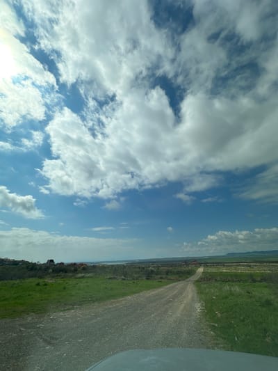 The image shows a dirt road in a rural area near Kosharitsa, Bulgaria. The road is bordered by green fields and a line of trees. The sky is blue with scattered white clouds. A portion of a vehicle is visible on the right side of the frame. In the distance, a few buildings and what appears to be a fence line can be seen.
