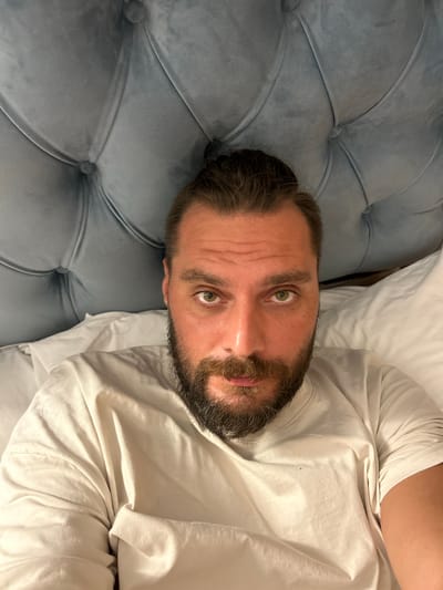 A close-up, vertically oriented, eye-level shot shows a man lying on a tufted, light blue surface that appears to be upholstered furniture. He is wearing a white t-shirt. His head is tilted to his left, and he is looking directly at the camera with a neutral expression. His eyes are green, and he has a short, dark beard and mustache. The lighting suggests it could be daytime indoors, possibly in a bedroom or living area. The setting appears to be in Kadewatta, Sri Lanka, though there are no specific visual cues to confirm this location within the image itself.