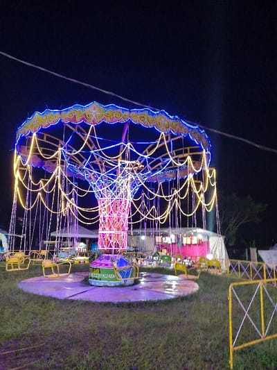 This image depicts a nighttime outdoor scene in An Khe Ward, Vietnam, centered on a stationary, illuminated swing ride (Chair-O-Planes type). The ride features a central pillar wrapped in pink and purple string lights, supporting a large circular canopy outlined with blue LED strips and draped with numerous yellow string lights. Below the canopy, multiple empty yellow swing chairs hang from chains. The ride rests on a circular, wet concrete platform situated on grassy ground. The text "AN KHU DEVELOPMENT" is visible on the greenish-blue base of the central column. In the background, white tent structures are visible, one with internal pink drapes illuminated by lights. Yellow metal fencing with cross-bar patterns demarcates the area on the right. The sky is dark, indicating night, with a faint diagonal wire or line visible in the upper left quadrant. No individuals or discernible activities are present.
