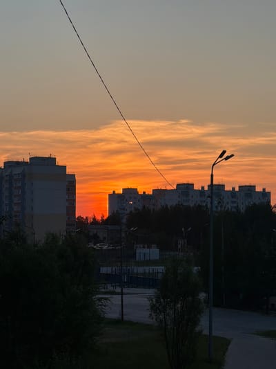 The image captures a sunset over Pyt'-Yakh, Russia. In the foreground, a dark silhouette of trees obscures the lower portion of the scene. A power line diagonally bisects the sky. In the middle ground, several multi-story apartment buildings, typical of Soviet-era architecture, are visible. The sky is dominated by vibrant orange and red hues, indicating the setting sun. A street lamp stands prominently in the mid-right of the frame. The background features more apartment buildings and dense foliage, with a road visible in the lower right corner.