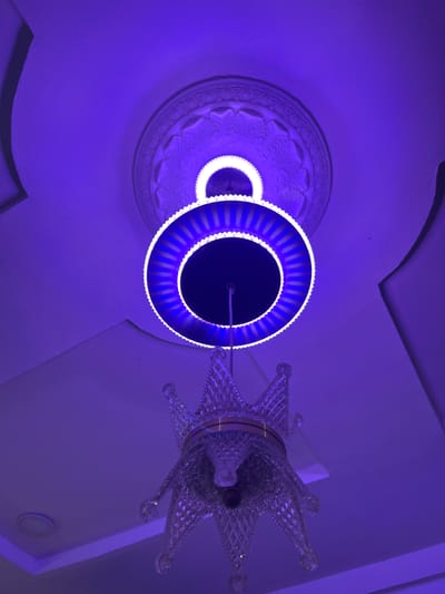 The image captures a close-up, low-angle view of a ceiling with ornate decorative molding and a modern chandelier. The lighting is predominantly a vibrant purple hue, suggesting artificial illumination. The chandelier itself is a striking object, featuring a circular LED light fixture with blue and white accents, surrounding a dark central area. To the right of the LED ring, a portion of a decorative crown-like structure made of clear, faceted material is visible. This crown element appears to be part of the chandelier, adding a touch of regal ornamentation. In the background, a section of the ceiling shows detailed plasterwork in a circular pattern, also bathed in the purple light. The overall scene creates an atmospheric and somewhat dramatic effect due to the intense color. The setting appears to be indoors, likely a room with a decorative ceiling. No people are visible, and the focus is entirely on the architectural details and the illuminated chandelier. The location context provided is Takau, Nigeria, but there are no specific visual cues within the image to confirm this. The time of day and weather are indeterminate due to the indoor setting and artificial lighting. No text is visible in the image.