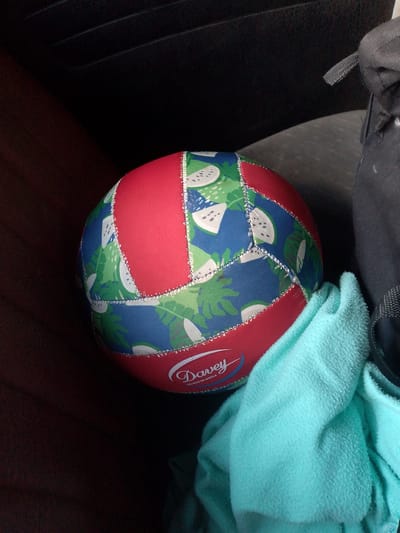 A close-up, slightly overhead shot shows a colorful volleyball with a pattern of green palm leaves and white slices of fruit on a blue background, interspersed with red panels. The volleyball has "Davey" and "CALIDAD DE MARCA" printed in white on a red section. It rests on a textured, dark brown surface, possibly leather, which could be a car seat. To the right of the volleyball, a light turquoise towel is draped over it and extends out of the frame. A dark gray bag or backpack is visible behind the towel and to the right of the ball. The background is dark and indistinct, suggesting an interior setting with low lighting, possibly the interior of a vehicle in El Maco, Venezuela.