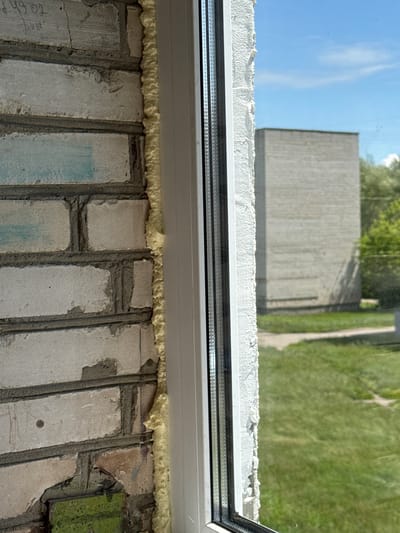 The image captures a close-up view of a window frame being installed against a brick wall. The left side of the frame is bordered by rough, light-colored bricks, some of which appear to have faint blue markings and possibly some graffiti. Expanding foam insulation is visible between the brickwork and the window frame, filling the gap. The window frame itself is white, with a visible inner metal reinforcement.
Through the window, a grassy area and a concrete path are visible. In the background, a large, light-colored rectangular building with a rough texture stands under a clear blue sky with a few scattered clouds. Trees can be seen behind this building, suggesting an outdoor environment. The scene appears to be in a residential or industrial area, possibly under construction or renovation. The overall impression is one of an urban or semi-urban setting on a bright, sunny day. Given the context provided for "Яромінскі сельскі Савет, Belarus", this scene likely depicts a window installation at a building within this administrative region.