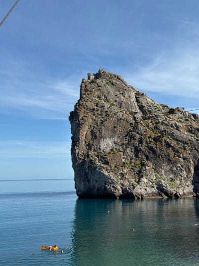 A towering, rugged cliff face juts out into the clear blue water of a body of water, possibly the sea. The rock is weathered and textured, with patches of green vegetation clinging to its surface. A metal railing and a series of steps are visible on the side of the cliff, suggesting a pathway or viewing platform. The sky above is a vibrant blue, with wispy white clouds.
In the water near the base of the cliff, a person is swimming, their head and shoulders visible above the surface. Further out, another person is floating on an orange inflatable raft, holding a ball. The water is calm and reflects the sky, with a slight ripple visible on its surface. The scene appears to be set on a sunny day, likely in the late morning or early afternoon, given the bright light and clear sky.
The location appears to be a coastal area, possibly with a dramatic drop from the land into the water. The presence of swimmers and a recreational raft suggests a popular spot for outdoor activities. The cliff face is the dominant feature of the scene, emphasizing its natural grandeur.