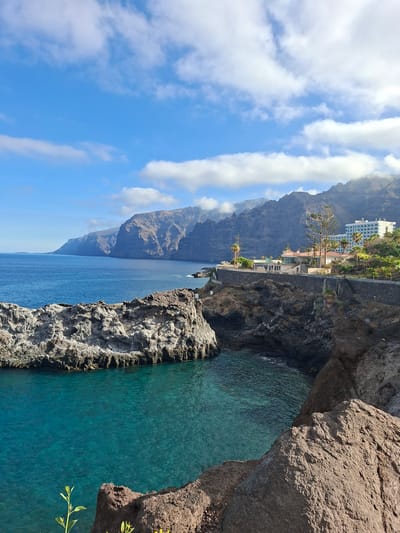 The image captures a scenic coastal view in Santiago del Teide, Spain. In the foreground, rugged, dark brown volcanic rock formations jut out into the vibrant, turquoise water of a cove. These rocks are textured and rough, with some patches of lighter-colored, weathered rock visible on the larger formation to the left.
Beyond the immediate rocky shore, the deep blue ocean extends towards a dramatic coastline characterized by towering, sheer cliffs. These cliffs, a dark gray and brown hue, rise majestically from the sea and stretch into the distance, creating a imposing backdrop. The sunlight illuminates parts of the cliff faces, highlighting their rugged texture and vertical striations.
Along the cliff edge, a paved pathway runs, separating the sea from a developed area. On this elevated platform, several buildings are visible. One prominent structure is a large, white hotel or apartment complex with multiple floors and numerous windows, positioned to offer views of the ocean. Adjacent to it, a smaller, earth-toned building with a distinctive red roof and arched windows appears to be a restaurant or other establishment, surrounded by greenery and palm trees.
The sky above is a bright, clear blue, punctuated by scattered white, fluffy clouds that cast soft shadows on the landscape below. The overall impression is one of a beautiful, sun-drenched coastal landscape with a blend of natural geological formations and man-made structures. There are no people or animals clearly visible in the image.