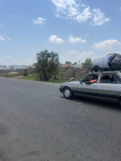 This media file captures a scene on a road in Wai A, Nigeria. The primary object is a light-colored sedan, overloaded with luggage strapped to its roof. The car is in motion, indicated by the blur of the asphalt road. A man, wearing an orange shirt, is visible driving the car, with his left arm on the steering wheel.
The setting appears to be a rural or semi-urban area, with houses and trees lining the side of the road. The architecture of the houses suggests a local style, with some appearing to be made of brick and others of corrugated metal. A prominent palm tree with distinctive fronds is visible to the left of the car. The sky is bright blue with scattered white clouds, suggesting it is daytime and the weather is fair. The overall impression is one of travel, possibly a long journey given the amount of luggage. There is no visible text in the media file.