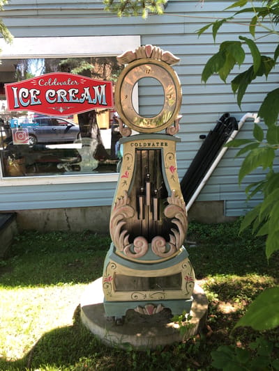 In the city of Severn, Canada, a weathered, whimsical grandfather clock-like structure stands on a concrete base, positioned outdoors beside a building with light blue horizontal siding. The clock features a large, circular opening at the top, with numbers 12, 3, and 6 visible. Decorative elements in shades of pink, yellow, and blue adorn its body, which houses chimes and appears to be a repurposed or artistic creation. To the left, a storefront window displays a prominent red sign with white lettering that reads "Coldwater ICE CREAM." Inside the window, a grey car is visible, along with reflections of trees and a sticker that appears to be an Instagram logo and text "Like Us on Facebook." The ground around the clock is covered in green grass and some small weeds, with dappled sunlight casting shadows. In the background, black pipes and a white pipe are leaning against the building. Lush green foliage from overhead branches partially obscures the top right portion of the image.