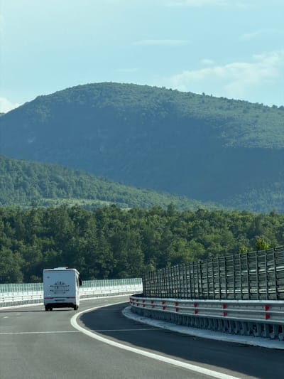 An outdoor, eye-level, medium shot captures a white camper van driving on a paved highway curving to the right. The camper van is in the left lane, facing away from the viewer. It has a license plate that reads "PU 7413 BG". A tall, dark, sound barrier fence runs along the right side of the highway, curving with the road. A metal guardrail with red reflectors is visible in front of the sound barrier. The highway is lined with white barriers. In the background, a lush, green, tree-covered mountain rises up against a hazy, light blue sky. The overall scene depicts a scenic drive through a mountainous region.
