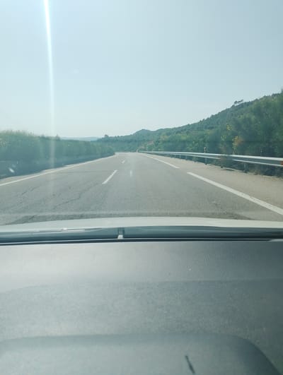 The image shows a view from inside a car driving on a two-lane highway. The road is asphalt with white lane markings. To the left and right of the road, there are green bushes and trees, with a hillside covered in more vegetation in the distance. The sky is clear and bright, with a strong lens flare from the sun visible on the left side. The dashboard of the car is visible in the foreground. The location is Abia de la Obispalía, Spain.