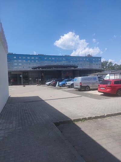 This media file captures an outdoor scene on a sunny day, likely in Chemnitz, Germany. The prominent subject is a light blue, multi-story building that appears to be an apartment complex or office building, with many uniformly spaced windows. In front of this building is a commercial area with glass-fronted shops, one of which has a "SALE %" sign visible.
Several vehicles are parked in the lot in front of the shops, including a silver van with "Automatiktüren GU" (Automatic Doors GU) and other text in German printed on its side, a red station wagon, and a blue SUV. A white truck is also partially visible. The ground is paved with interlocking bricks and concrete. The sky is a bright blue with scattered white clouds, suggesting fair weather and daytime. The overall impression is a mundane, everyday scene in a commercial or residential area. No people are clearly visible.
