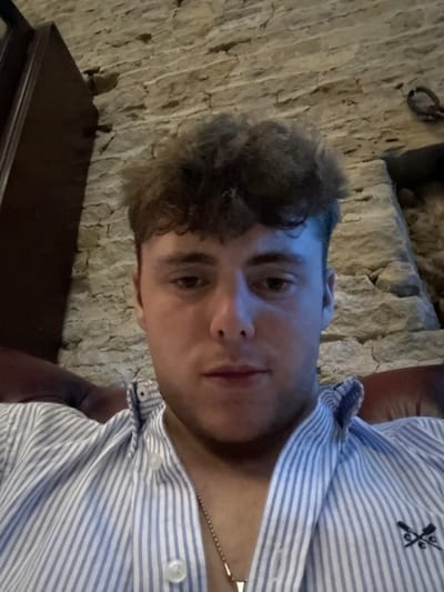 A young man with curly brown hair is posing for a selfie. He's wearing a blue and white striped shirt with a logo on the collar, and a thin gold chain around his neck. The background shows a textured stone wall behind him and a dark wooden table beneath him. The lighting appears to be dim and somewhat artificial, creating shadows on his face and the surrounding environment.