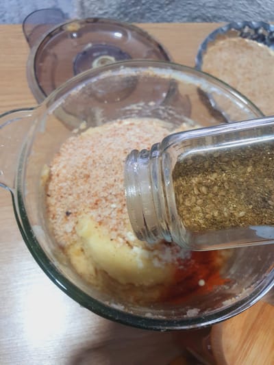 A close-up shot captures a glass bowl filled with a mixture of ingredients, including what appears to be mashed potatoes, breadcrumbs, and red spices. A glass spice jar, tilted at an angle, is pouring more spice into the bowl, its contents a blend of small seeds and dried herbs. The scene is set on a wooden surface, with blurred background elements suggesting a kitchen environment, possibly including a food processor lid and another dish containing breadcrumbs. The lighting is bright, highlighting the textures and colors of the food ingredients.