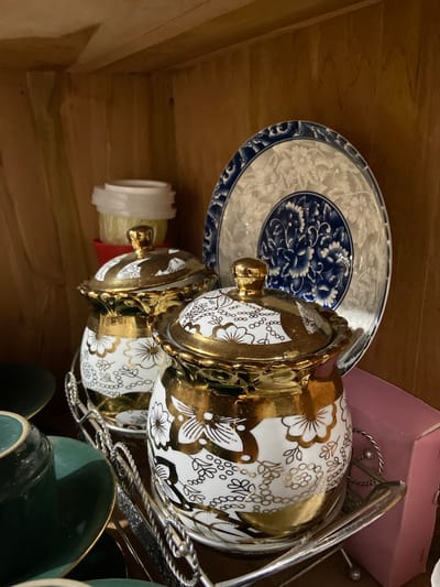 A close-up, eye-level shot shows a collection of vintage porcelain items displayed on a wooden shelf. In the foreground, two lidded canisters with ornate gold floral and vine patterns on a white background are prominently featured. They are placed on a silver metal rack with twisted handles. To the left, a stack of green ceramic cups and saucers, with gold trim, are partially visible. Behind the canisters, a large, decorative plate with intricate blue and white floral patterns stands upright. The background consists of the wooden shelves and a glimpse of a pink box on the right. The lighting is dim, creating shadows and highlighting the reflective surfaces of the gold and silver. The scene suggests an indoor display, likely a home or shop, showcasing decorative dishware. There are no people or animals present in the image. The image does not contain any information about the location in Margorejo, Indonesia.
