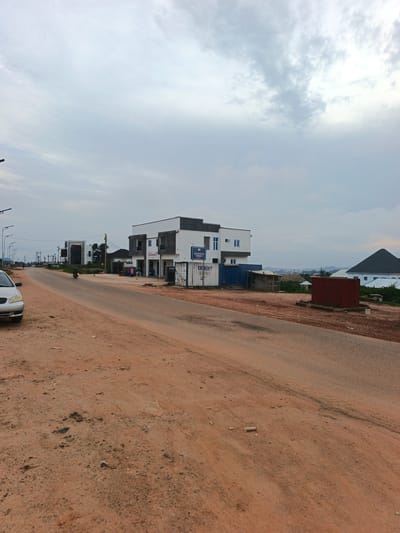 A dirt road leads past several buildings and a few shipping containers under a cloudy sky. The road is mostly reddish-brown dirt with some gravel and patches of dry grass. On the left side of the road, a silver sedan is parked. Further down the road, a motorcycle carrying a rider is moving away from the camera. On the right side of the road, there is a modern two-story building with white and grey facades. A sign on the building reads "PHARMACY." Next to this building, there are two shipping containers, one blue and one white. The white container has text that includes "DANGOTE CEMENT" and "CEMENT DEPOT." There is also a small, simple wooden shelter next to the containers. In the background, more buildings and structures can be seen, along with a dark, conical-roofed building. The overall impression is a developing area with commercial activity. The time of day appears to be late afternoon or early evening, given the lighting. The weather is overcast.