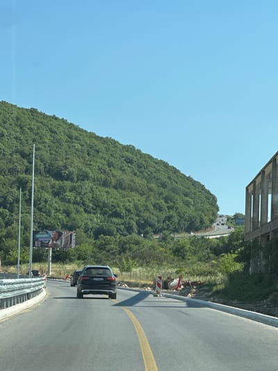 The image captures a winding road in Prijeradi, Montenegro, on a clear, sunny day. A black SUV is driving on the road, followed by another car. To the left of the road, a lush green hillside is covered in dense trees. In the distance, more cars are seen navigating the curves of the road. On the right side of the road, a partially constructed concrete building stands with its skeletal framework exposed. Construction barriers and materials are visible along the roadside, indicating ongoing work. A billboard advertising Nescafe is also present on the left side, positioned near tall poles. The road itself is paved and marked with a yellow line down the center, suggesting a moderately trafficked route. The environment appears to be semi-rural, with the natural landscape dominating the scene.