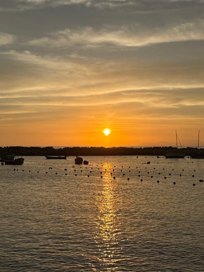 The image captures a sunset over the water in Chorrillos, Peru. The sun is positioned on the horizon, casting a bright orange glow across the sky and reflecting on the water's surface. The sky features elongated cloud formations. A line of what appears to be a rocky coastline or breakwater divides the sky from the water. Several buoys are visible in the water. A mast of a boat is silhouetted against the bright sky.