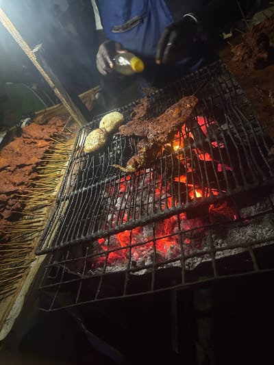 The image depicts a nighttime scene of an outdoor grilling operation, likely in Galadimawa, Nigeria. The central activity involves the preparation of food over a vibrant charcoal fire.
On the left side of the frame, a person's dark-skinned hand, clad in a blue sleeve, is visible holding a small, yellow-capped bottle, possibly containing oil or a spice blend for basting. The hand is positioned near a robust metal grill grate, under which bright red, glowing charcoal embers provide intense heat and the primary light source for the scene.
Cooking on the grill are several skewers of seasoned meat, typical of "suya," a popular West African grilled dish. Their surfaces appear well-cooked and rich brown. Additionally, two lighter-colored, rounded food items, possibly potatoes or similar accompaniments, are roasting alongside the meat. Below the main grill, a wooden surface holds a substantial quantity of raw, reddish-brown marinated meat skewers, arranged on sticks, indicating ongoing preparation or a busy stall.
Smoke subtly rises from the cooking area, illuminated by the hot coals. The rustic setup suggests a street food vendor or an informal outdoor eatery, operating in the dark, with the fire's glow creating a warm and inviting atmosphere. No visible text is present in the image.