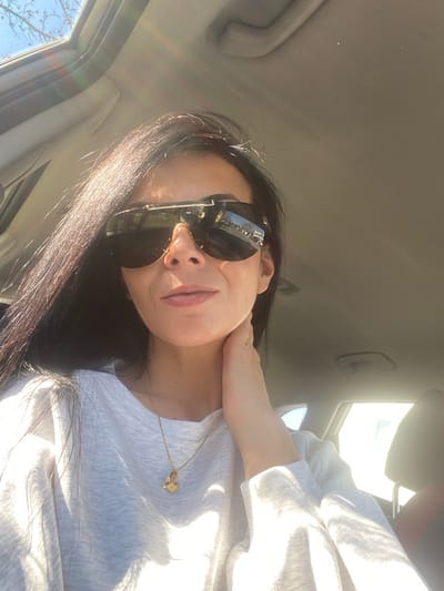 The image displays a female individual seated inside a vehicle, identified as being in Bensheim, Germany. The subject has dark, long hair and is wearing large, reflective sunglasses with gold-colored accents on the frame. She wears a light-colored, possibly grey or white, long-sleeved top and a gold-colored necklace featuring a heart-shaped pendant. Her right hand is raised, with fingers touching the area near her chin and neck. The interior of the vehicle includes a light-colored headliner and a partially open sunroof, through which a clear blue sky and bare tree branches are visible. Direct sunlight strongly illuminates the scene, causing lens flares and pronounced highlights, suggesting a bright outdoor environment. Reflections visible on the sunglasses lenses show an exterior scene consisting of bare trees and a paved area, consistent with a street or parking lot.