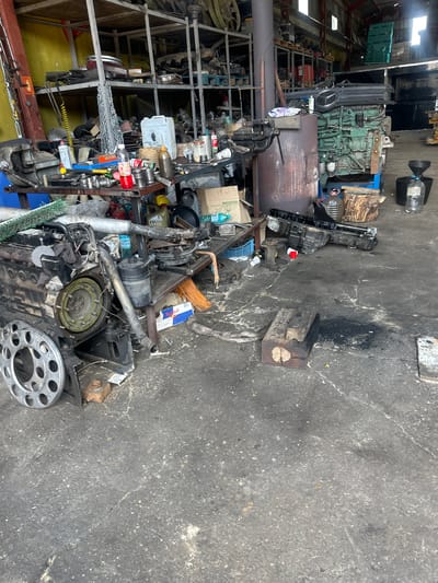 The image depicts a cluttered workshop environment, likely a mechanic's garage, filled with various automotive parts and tools. In the foreground, a large engine block is prominently displayed, surrounded by smaller engine components, a metal wheel rim, and what appears to be a wooden block. Various containers, bottles, and cans are scattered on a workbench and the floor. In the background, shelves are stacked with more parts, and a large green engine, possibly for a truck or industrial vehicle, is visible. The overall impression is one of disarray and ongoing mechanical work. No people are visible in the image. The scene is set within a building with a concrete floor and exposed structural elements.