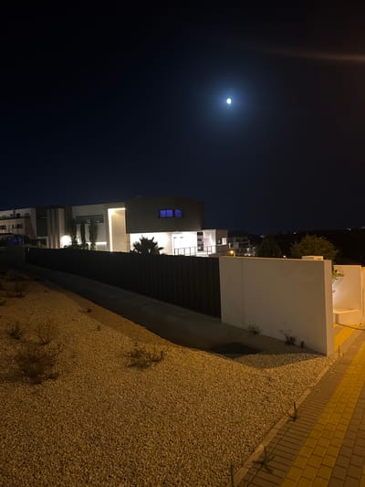 The image shows a modern, multi-story building complex at night in Finestrat, Spain. The sky is dark blue with a bright full moon visible in the upper right quadrant. The building features clean lines, geometric shapes, and large windows, some of which are illuminated with blue light. A dark, vertical fence runs along the front of the property, separating it from a sloped area covered in light-colored gravel and sparse vegetation. To the right of the fence, a white wall is visible, adjacent to a paved walkway with yellow and grey brickwork. The scene is illuminated by artificial lights from within the buildings and from streetlights, casting shadows on the gravel. No people are visible in the image.