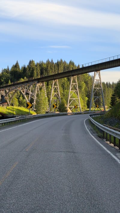 A paved road curves through a scenic landscape, with a large, multi-span bridge dominating the upper portion of the image. The bridge, an impressive structure of steel girders and supports, spans over a wooded area. The road itself is asphalt with visible yellow lane markings.
Flanking the road are metal guardrails, with the one on the right showing some weathering and posts. Yellow chevron signs, indicating a curve in the road, are strategically placed along the side of the road, drawing attention to the bend. In the background, a dense forest of evergreen trees stretches across the horizon, their dark green foliage contrasting with the bright blue sky. The sky is partly cloudy, with wispy white clouds scattered across it.
The sunlight suggests it is either early morning or late afternoon, casting a warm glow on the scene. The overall impression is one of a serene and natural environment, with the man-made bridge being a prominent, yet integrated, feature of the landscape. There is a small sign with Norwegian text visible on the left side of the road near the bridge structure, partially obscured by grass. The text on the sign appears to read "OVERNATTING", which translates to "ACCOMMODATION" in English. No people are visible in the image.