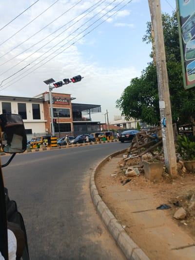 The image depicts a street scene in Kabala, Nigeria, from the perspective of someone riding in a vehicle (possibly a tricycle, judging by the partially visible mirror and frame). The road curves to the left. On the left side of the road is a two-story building with a sign that reads "A-CLASS RESTAURANT." A solar-powered traffic light is visible above the restaurant. Several vehicles, including cars and yellow and green tricycles (Keke Napeps), are parked or moving along the street. On the right side of the road is a curb, followed by a dirt and concrete sidewalk with trash and debris. A utility pole is present on the right edge of the frame, partially obscured by foliage and what appears to be makeshift wooden support. The sky is partly cloudy.