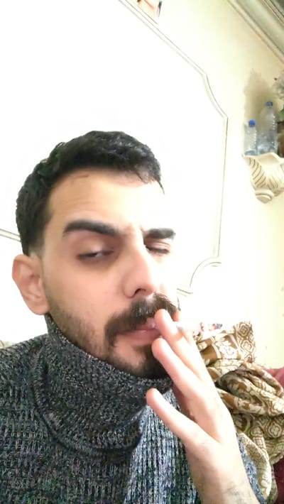 The image displays a male individual, seen from the upper chest upwards, in an indoor setting within Al-Qaboun Municipality, Syria. He has dark, short hair, a mustache, and a beard. His eyes are partially closed or directed downwards. He is wearing a dark, textured turtleneck sweater. His right hand, positioned near his mouth, holds a lit cigarette between his thumb and forefinger. The background consists of a light-colored wall featuring a decorative architectural molding. To the right, a wall-mounted shelf holds two plastic water bottles, one larger than the other. Below the shelf and to the right of the individual's shoulder, a pile of patterned brown and beige fabric is visible.
