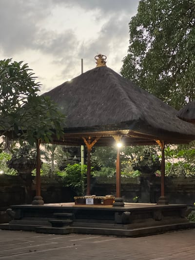 The image shows an outdoor scene at dusk, featuring a traditional Balinese gazebo with a thatched roof. The gazebo is elevated on a stone platform and supported by wooden pillars. A golden crown-like ornament is visible on the peak of the thatched roof. Inside the gazebo, a small sign with Balinese text reads "Sumbangan Dana Punia." The surrounding area is lush with greenery, including trees and bushes. In the background, glimpses of stone temple structures can be seen, suggesting the location is a temple or a culturally significant site within Dawan, Indonesia. The lighting is dim, with a single artificial light source illuminating the interior of the gazebo. The sky is overcast with a pale, diffused light. There are no people or animals present in the frame.