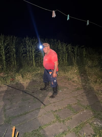 A man stands in a paved area at night, illuminated by a headlamp. He is wearing a red shirt, blue pants with orange accents, and black boots. A cornfield is visible behind him, and a clothesline with clothespins is strung across the top of the image. The location is in Tandzia, Georgia.