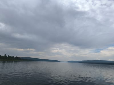 The image shows a wide, calm river under a cloudy sky. The river occupies the foreground and midground of the image, with ripples visible on its surface, indicating a gentle current. On the left bank, a dense line of green trees stretches along a low, undulating hill. The right bank is also characterized by a similar low, forested hill that recedes into the distance. The sky is filled with heavy, grey clouds, with some lighter patches suggesting diffused sunlight. The overall atmosphere is somber and natural. The scene is consistent with a view from the Angara River in the vicinity of Boguchany, Russia, a region known for its wide rivers and forested landscapes. No people or specific man-made objects are discernible in the image, aside from potentially distant power line pylons on the horizon.