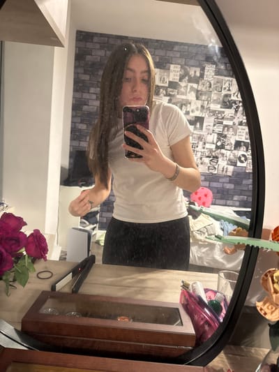A person stands in front of a round mirror, holding a phone to take a selfie. They are wearing a white t-shirt and dark pants. The background includes a wall decorated with many small pictures, a bed, and various items on a surface including red roses, a hair straightener, and a wooden display case. The reflection in the mirror reveals a casual indoor setting.