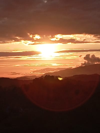 A picturesque sunset unfolds over a mountainous landscape in Bagazhvyashta, Abkhazia. The sky is ablaze with warm hues of orange and yellow, with streaks of sunlight piercing through the clouds. Silhouetted against this vibrant backdrop are layers of mountains, their peaks fading into the distance. The foreground is dominated by a dark, forested area, its details obscured by the fading light. A circular lens flare, characteristic of the sunset's intensity, is visible in the lower portion of the image, adding a touch of atmospheric effect to the scene. The overall impression is one of serene natural beauty, capturing the tranquil end of day in this Abkhazian locale.