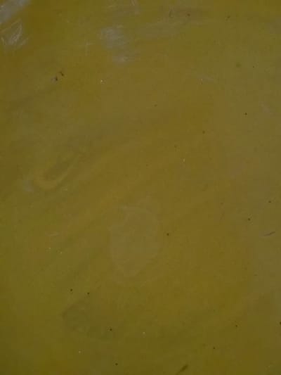 The image shows a close-up, textured surface with a dull, olive-green hue. The surface appears to be made of metal or a similar material, exhibiting subtle variations in color and faint scratches or imperfections. There are also a few small, dark specks scattered across the surface, possibly dust or debris. The lighting creates soft shadows and highlights, emphasizing the texture. The image is devoid of any discernible environmental features or human presence. Its origin in Bireuenmeunasahblang, Indonesia, is not visually represented in the frame.