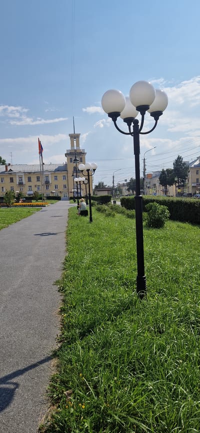 The image shows a paved walkway on the left, bordered by a lush green lawn on the right. Multiple streetlights with spherical white globes are positioned along the walkway. In the background, a light yellow building with a prominent tower stands out. Two flags, one red and one orange, are visible on flagpoles in front of the building. Several people are seated on benches or standing, engaging in various activities. The environment appears to be an urban park or public space on a sunny day, with a clear blue sky overhead. The scene is located in Miass, Russia.