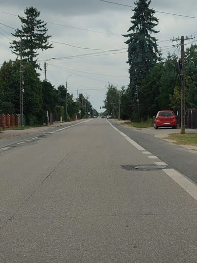 This is a medium-shot, eye-level, vertical photograph of a two-lane asphalt road in Łomna, Poland. The road runs from the foreground into the distance, with dense trees lining both sides. Power lines and streetlights are visible overhead. On the right side of the road, a red hatchback car is parked facing away from the camera. In the distance, a few other cars are visible on the road. The sky is overcast. The scene appears to be a suburban or rural area.