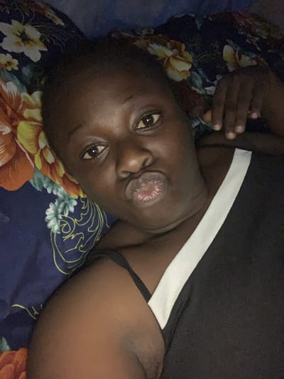 A person with dark skin is depicted in a close-up, upward-angled portrait, lying down and looking directly at the camera. Their head is tilted to the side, resting on a patterned bedspread. The individual's lips are pursed in a distinctive "duck face" or "fish-gaping" expression, revealing the inner surface of the lower lip. Their eyes are open, reflecting a light source, and convey a direct gaze.
The person is wearing a dark-colored top, possibly black, featuring a prominent white horizontal stripe across the upper chest or shoulder. A thin, dark strap, possibly from an undergarment or another layer of clothing, is also visible. The background is composed of a dark blue sheet or blanket adorned with large, vibrant floral patterns in orange, yellow, and light blue, interspersed with light blue abstract swirling lines.
The lighting in the image is dim, suggesting it was captured indoors during low-light conditions, likely at night or in the evening. The overall ambiance is relaxed and playful, indicating the person is posing for a photograph or selfie. There is no visible text within the image. The location context for this scene is Kukum, Nigeria.