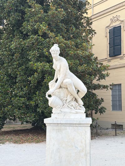 A white marble statue of a female figure pouring from a jug is positioned on a pedestal in a park in Parma, Italy. The statue depicts a woman in a seated position, leaning forward with her right arm extended, holding a jug. Her hair is styled in a bun. Behind the statue, a large, dense tree with dark green foliage dominates the background. To the right of the tree, a light yellow building with a dark-shuttered window and a smaller barred window is visible. The ground in front of the statue is covered with light-colored gravel, and fallen leaves are scattered beneath the tree. A low metal chain fence with posts is visible in the mid-ground. The statue is located in the Parco Ducale in Parma.