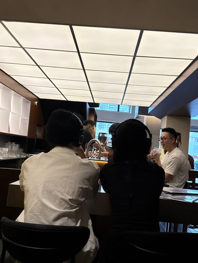The image shows a group of people in what appears to be an indoor setting, possibly a coworking space or a modern office. The orientation of the image is rotated 90 degrees clockwise.
In the foreground, a person wearing a white shirt and headphones is facing away from the camera, with their head tilted slightly down. Their hair is dark and short. To their left, another person wearing a black shirt and headphones is also facing away from the camera. Their dark hair is visible.
Further in the background, a person wearing glasses and a white t-shirt is looking towards the left of the frame, holding a clear plastic cup. Their expression is not clearly discernible due to the angle and lighting. Another person, wearing dark clothing, is partially visible behind them.
The environment features a large, illuminated panel system on the left side of the frame, with a grid pattern of light squares. The rest of the background is dimly lit, with some wooden elements and glimpses of windows showing an urban outdoor scene with buildings. There are also shelves and what looks like a chair in the foreground.
The overall mood seems focused and collaborative, with individuals engaged in activities that likely involve concentration, possibly related to work or study, given the headphones and the general atmosphere of a modern workspace. There are no visible signs of the time of day or weather conditions. The location context provided is Jing'an District, China, which aligns with the appearance of a contemporary urban environment. No text is visible in the image.