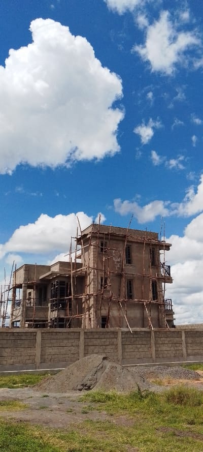 The image shows a partially constructed building in Pridelands, Kenya (latitude: -1.352, longitude: 36.976). The building is three stories tall and is made of concrete. It is surrounded by scaffolding. There is a concrete wall in front of the building, and a pile of dirt and gravel in front of the wall. The ground in front of the wall is covered in grass. The sky is blue with white clouds. The scene is located in a residential area. There is no sign of any people or animals. The image was taken in a clear, sunny day.  The scene is calm and serene.
